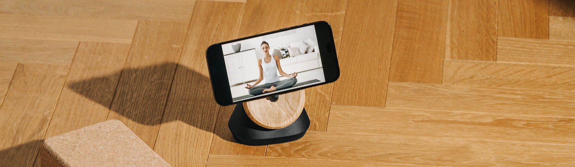 Smartphone mit Meditationsvideo auf einem Holzständer, daneben ein Korkblock auf Parkettboden