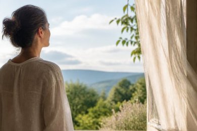 Eine Person steht im hellen Morgenlicht an einem offenen Fenster und blickt in eine weite grüne Landschaft.