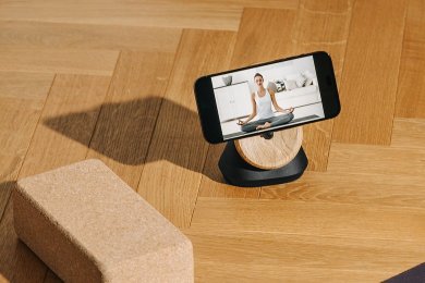 Smartphone mit Meditationsvideo auf einem Holzständer, daneben ein Korkblock auf Parkettboden